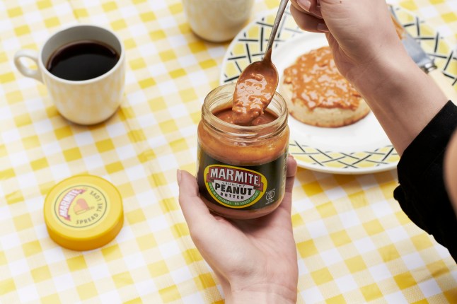 A jar of Marmite peanut butter with a spoonful being scooped out. In the background is a breakfast set up with a mug of coffee and a crumpet with Marmite Peanut butter on it