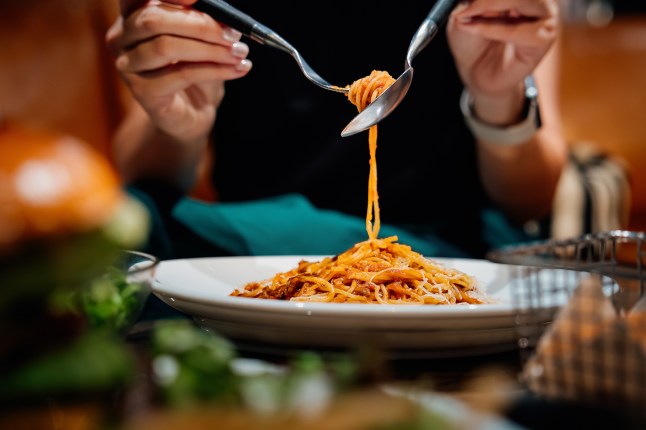 Close-up, mid-section of young woman sitting at dining table holding cutleries, twirling freshly served spaghetti bolognese and enjoying her meal in restaurant.